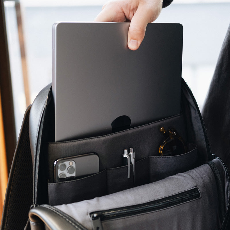 Mão inserindo um notebook em compartimento acolchoado da mochila Slim Workplace NW108, com bolso organizador contendo celular, canetas e óculos de sol.