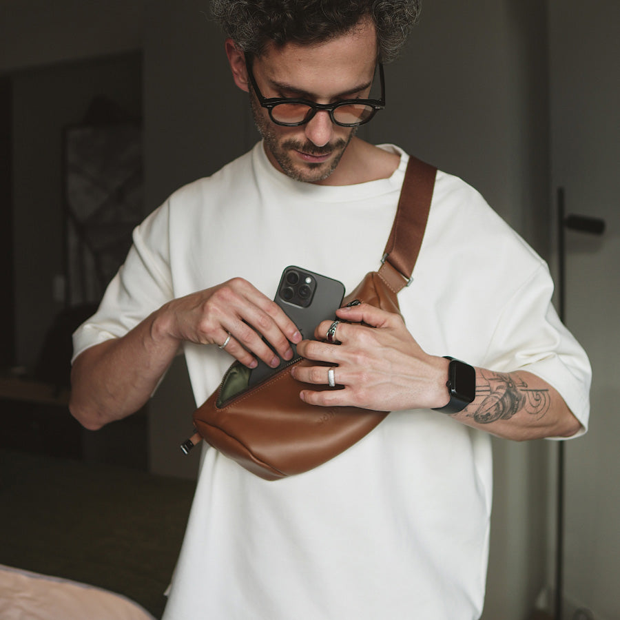 Homem utilizando a Sling Bag Pulse NW112 Nordweg de forma transversal no peito, guardando o celular no bolso principal da bolsa de couro caramelo.